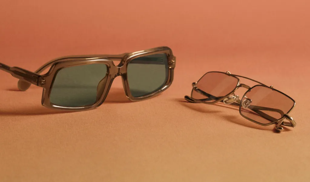 Dwie pary okularów Oliver Peoples leżące na ceglastym tle